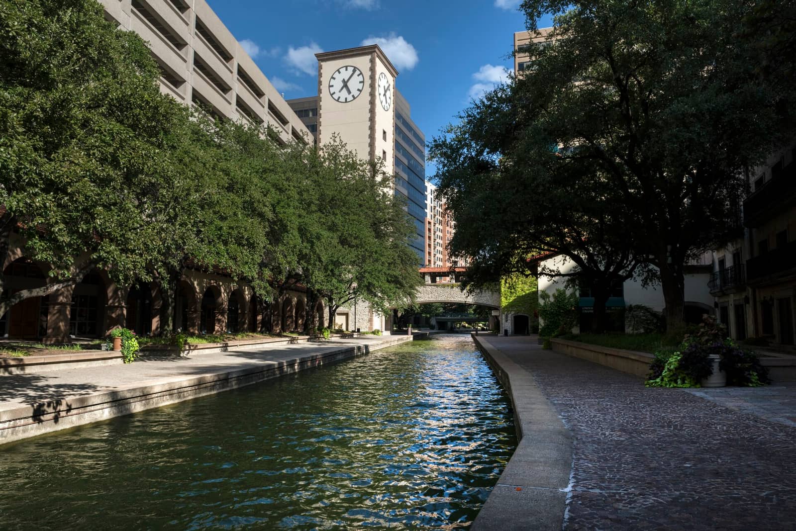 A quiet, tree-lined canal walkway with a clock tower and office buildings in the Las Colinas area of Irving, Texas